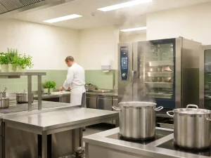 Chef in witte koksbuis werkt in professionele zorgkeuken met roestvrijstalen werkbladen, kookpotten en combi-steamer.